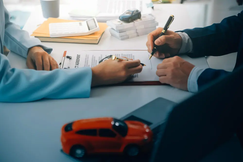 Two people signing insurance documents at a desk with a small red car model in front.