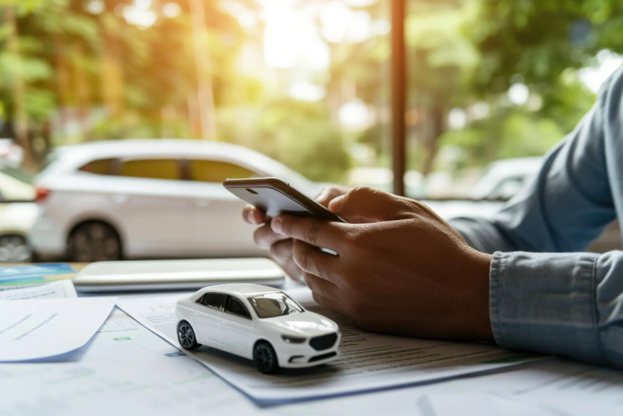 Person comparing vehicle coverage options on smartphone beside toy car models and insurance paperwork